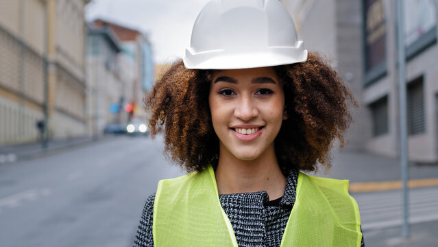 Female portrait worker profession close-up african american woman girl with curly hair civil engineer professional wearing safety helmet standing in city street and looking at camera smiles friendly
