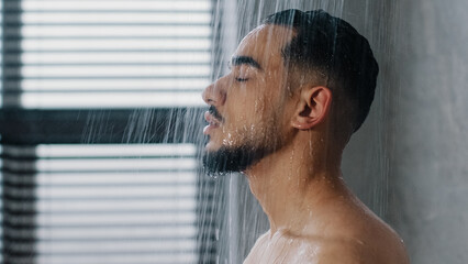 Side view indian relaxed man bearded guy naked male washes head hair face and beard in bathroom showering in at home in hotel after workout close up. Brunette rinsing shampoo and conditioner