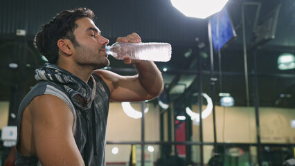 Side shot of Asian male bodybuilder is sitting on fitness bench and drinking water after tired from...