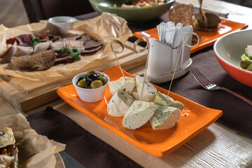 Cheese cubes with rosemary, olives and olive oil sauce in orange bowl. In the background is a set table.