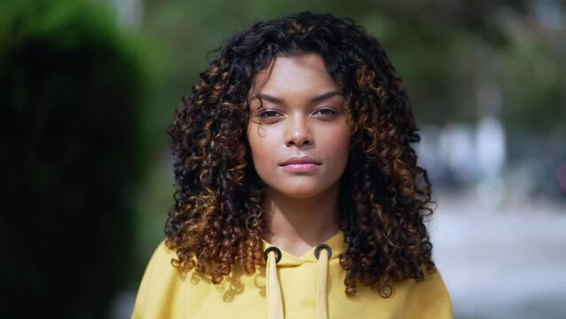 Portrait of a serious black hispanic latina girl with curly hair standing outside looking at camera. African American young millennial woman in 30s wearing yellow blouse
