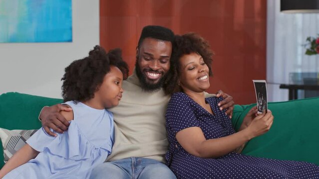 Happy African-American couple with daughter looking at sonogram picture sitting together on couch