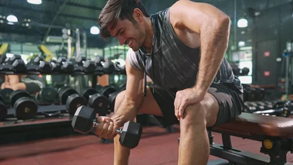 Fotobehang Asian male bodybuilder is sitting on bench and doing one-arm bicep curl with dumbbell exercise in fitness gym. Athlete man with motivation concentrate on lifting dumbbell to maintain bicep muscles. © Benixs