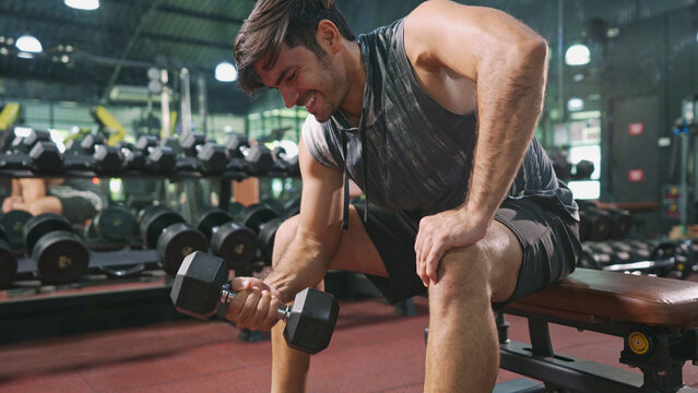 Asian male bodybuilder is sitting on bench and doing one-arm bicep curl with dumbbell exercise in fitness gym. Athlete man with motivation concentrate on lifting dumbbell to maintain bicep muscles.