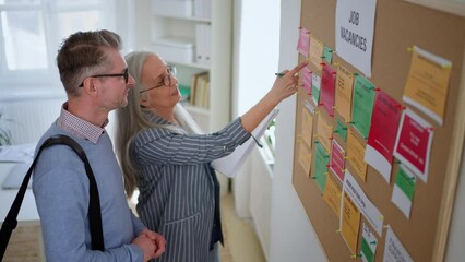 Recrutiment agency employee standing in front of employment noticeboard and helping mature man search for a job.
