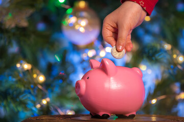 Children's hands put a coin in a pink piggy bank on a New Year's bokeh background. The concept of investment in the future of the child, savings, financial planning, fulfillment of desires