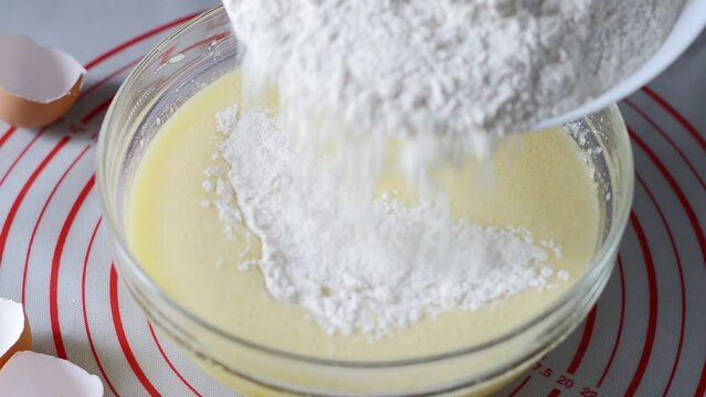 Woman pours flour into a bowl of batter and mixes the ingredients with a whisk.