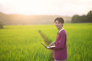 Asian men farmer stand use laptop at the gold rice field to take care of her rice. young own business start up farm. communication network on computer technology.