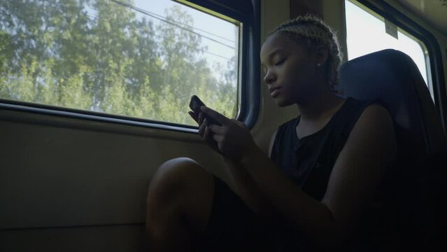 A young woman sits at the window in the train, writes a message on a mobile phone in her hands, trees outside the window, sun, summer. Slow motion 4k footage