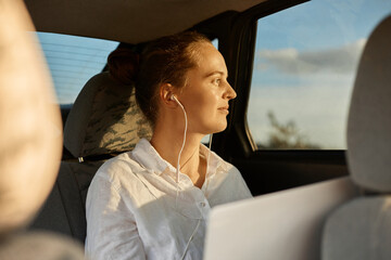 Side view portrait of beautiful woman freelancer wearing white shirt sitting on back seat of car and working with laptop, listening music and looking at vehicle window.