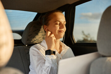 Photo of calm relaxed adorable woman wearing white shirt sitting on back seat of car and working...