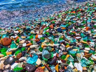 Natural polish textured sea glass and stones on the seashore. Azure clear sea water with waves. Green, blue shiny glass with multi-colored sea pebbles close-up. Beach summer background.