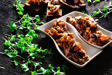 Appetizer of fried eggplant with prunes in unusual plates, a lot of microgreens scattered around, on a black textured background, delicious food