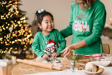 A Chinese mixed-race mother and a little daughter in green pajamas together prepare festive cookies from dough for the Christmas holiday in a decorated house in winter. Selective focus