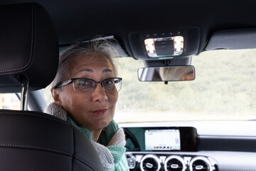 A gray-haired lady sits in the driver's seat of a car and looks around at the passengers.