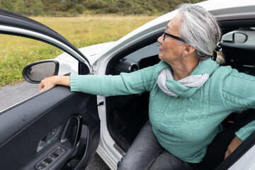 A gray-haired lady sits in the driver's seat in a car with an open door and looks away