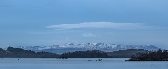 Beautiful landscape image of Loch Lomond and snowcapped mountain range in distance viewed from small village of Luss