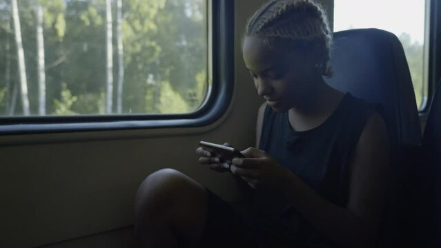 A young woman sits by the window in the train and plays a game on her mobile phone. Outside the window is summer, sun, trees. Slow motion 4k footage