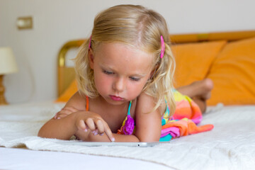 Little blond girl lay on bed and play tablet,toddler using internet with gadget,kid addicted to tablet and looking cartoons,3 year old child studiyng online with developmental games