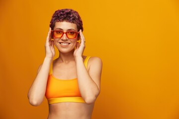 Young woman in a bright yellow fitness tracksuit with an open belly and sunglasses on an orange background smiling
