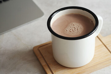 A mug of hot cocoa stands on a wooden stand on the table. Behind is a laptop
