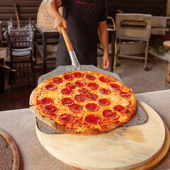 Pepperoni pizza on spatula held by pizza chef. Pizza oven in the background. Wooden board