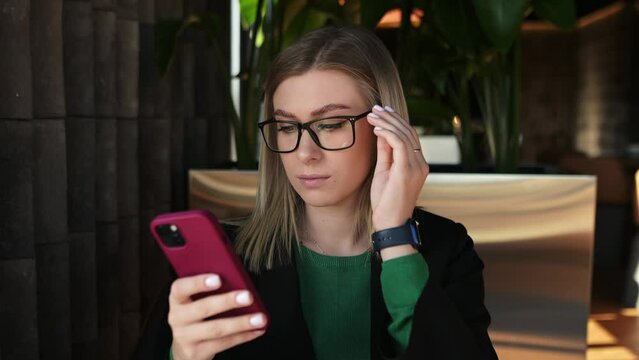 A young confident girl with a phone in her hand puts on glasses and looks confidently into the camera. Remote work, influencer, social media marketer