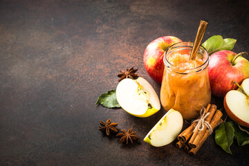 Apple jam with spices in glass jar and fresh apples on dark stone background.