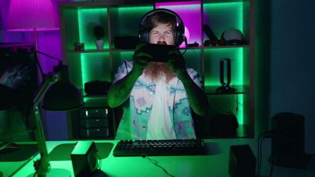Young redhead man streamer playing video game using smartphone at gaming room