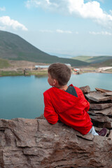 Fototapeta premium a teenager boy in a red jacket sits on the rocks high in the mountains and admires the natural beauty of the mountain landscape and the lake in the mountains.