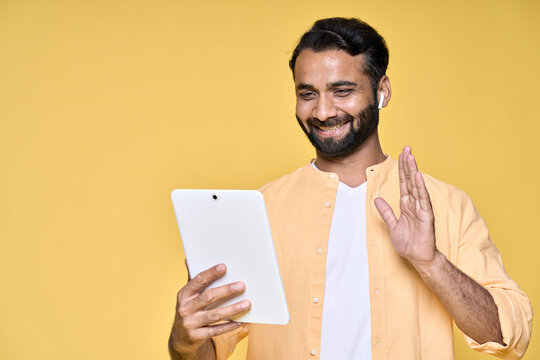 Happy indian man using digital tablet standing isolated on yellow background. Smiling ethnic guy wearing earbud waving hand having remote video call, remote online distance learning conference class.