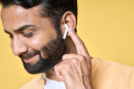 Happy smiling bearded indian man relaxing wearing earbud listening to music, business podcast or audio book in mobile application by earphone isolated on yellow background. Close up portrait.