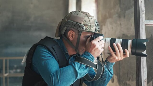 Close up of one man in helmet and bulletproof vest holding professional camera taking photos in war zone. Dangerous job. Male war journalist with digital camera working at place of action. War press