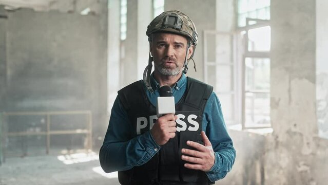Close up portrait of middle-aged Caucasian handsome correspondent reporting live from place of battle. Man journalist in war zone talking in microphone looking at camera. War press