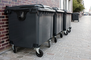 Several new gray plastic dumpster stand near the brick wall of the building, on a city street.