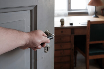 Hhand holds on to the handle on an open wooden door, through which one can see an office, an...