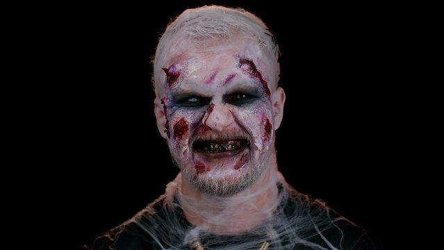 Sinister man with horrible scary Halloween zombie makeup in costume showing dirty teeth, looking ominous at camera trying to scare. Dead guy with wounded bloody scars face isolated on black background