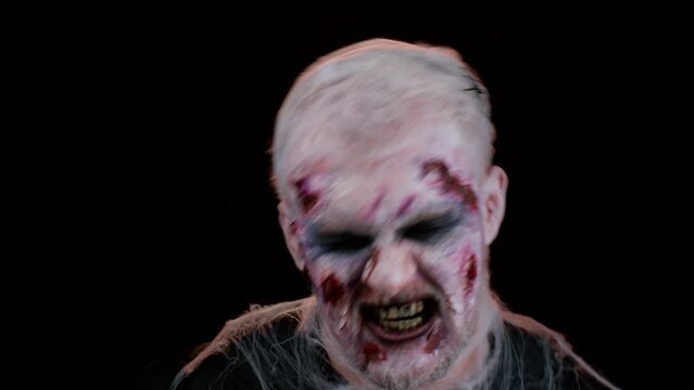 Zombie man with makeup with fake wounds scars and white contact lenses looking at camera and screaming, trying to scare in dark studio room. Sinister dead guy. Halloween, filming, staging concept