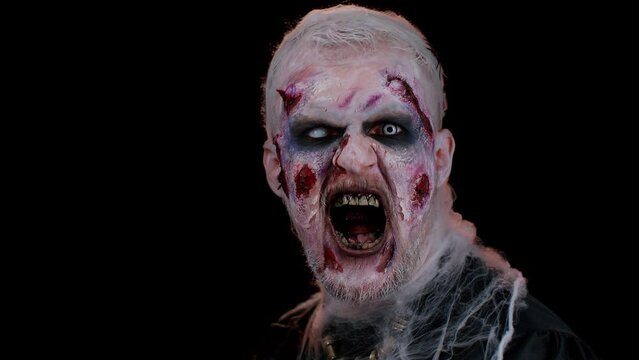 Zombie man with makeup with fake wounds scars and white contact lenses looking at camera and screaming, trying to scare in dark studio room. Sinister dead guy. Halloween, filming, staging concept