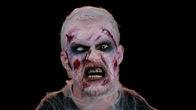 Unexpected appearance of sinister man in costume of Halloween crazy zombie with bloody wounded scars face trying to scare isolated on black room. Horror theme of cosplay wounded undead, beast, monster