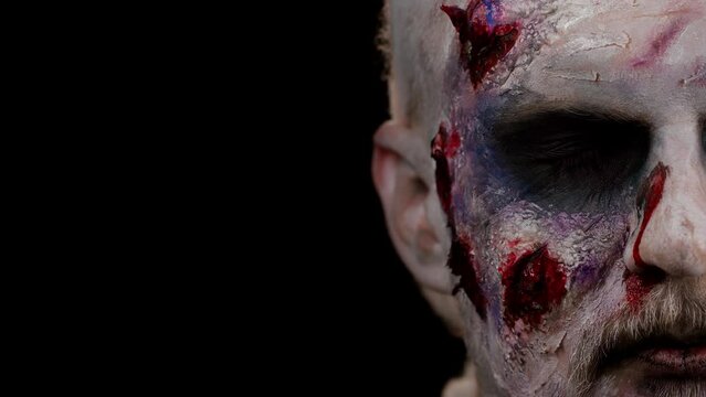 Close-up macro frightening man face with Halloween zombie bloody wounded makeup, trying to scare, face expressions. Horror theme. Sinister undead guy isolated on studio black background. Fashion art