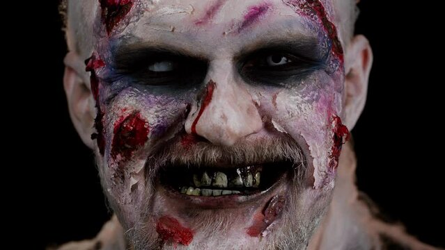 Close-up Halloween zombie man face makeup with wounds scars and white contact lenses looking at camera, trying to scare, showing tongue, dirty teeth in dark studio room background. Sinister dead guy