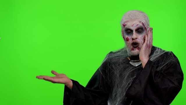 Zombie man with makeup with fake wounds scars showing thumbs up and pointing empty place, advertising area for commercial text logo, copy-space for goods promotion. Sinister Halloween ugly dead guy