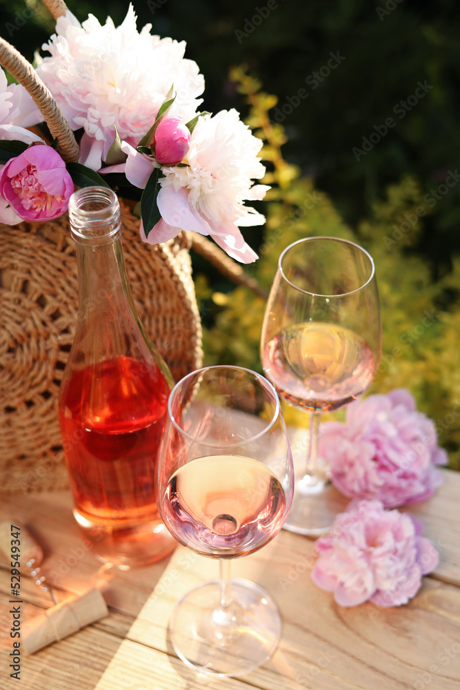 Sticker Bottle and glasses of rose wine near beautiful peonies on wooden table in garden