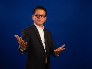 Portrait of smart middle-aged asian confident successful businessman wearing suit with welcoming gesture on blue background in studio, looking attractive and positive professional leadership