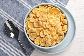 Bowl of tasty corn flakes and spoon on white wooden table, flat lay