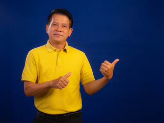 Portrait of middle-aged asian man in casual yellow T-shirt pointing finger in copy space isolated on blue background in studio, looking smart and attractive