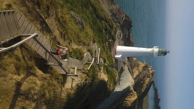 A woman jogs down a wooden boardwalk toward a white lighthouse in New Zealand. Vertical shot.