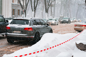 Many cars move down a snowy city's road in winter with red and white barrier tape on the side....