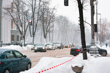 Many cars move down a snowy city's road in winter with red and white barrier tape on the side. Dirty. Snow. City. Winter. Street. Outdoor. Transportation. Blizzard. Heap. Climate. Weather. White